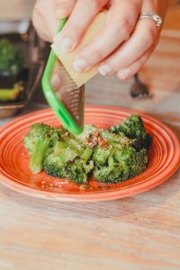 women shredding parmesan cheese on a small plate of cooked broccoli