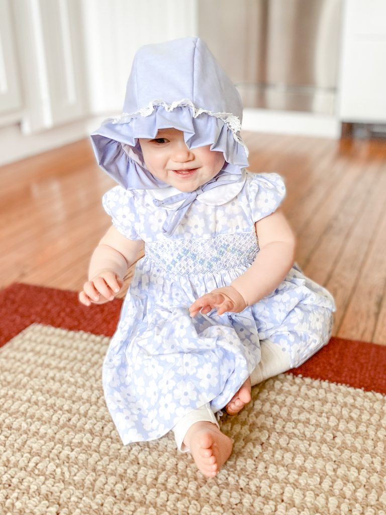 Revival Acres baby in blue dress in bonnet sitting on floor