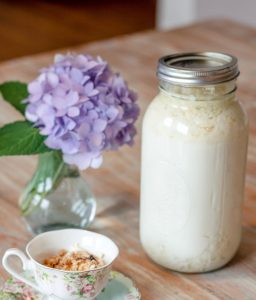 a jar of homemade kefir sitting on a tbale with a teacup and purple hydrangea flower